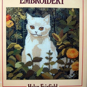May include: Book cover for "Counted-Thread Embroidery" by Helen Fairfield. The cover features a white cat sitting in a garden with green leaves and orange flowers.