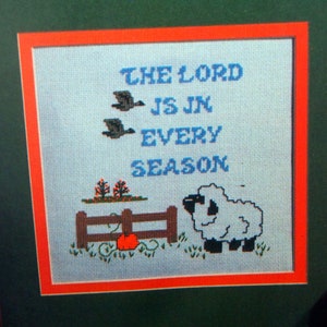 May include: Cross-stitch artwork in a wooden frame. The design features the text "THE LORD IS IN EVERY SEASON" in blue, with images of geese, a fence, a pumpkin, and a sheep. The artwork is framed with a green mat and an orange border.