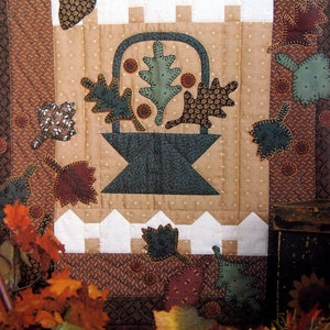 Pode incluir: Um cobertor marrom e branco com uma cesta de folhas no centro. O cobertor é feito de vários tecidos e tem um design de patchwork. A cesta é cercada por uma cerca branca de madeira.