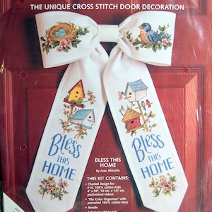 May include: Cross-stitch door decoration kit featuring a white bow with embroidered birdhouses, birds, and floral accents. The text "Bow Ties" and "Bless This Home" are displayed. Finished size is 11" x 17".