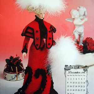 May include: A crocheted doll wearing a red dress with black trim and a white feathered hat. The doll is holding a white feathered boa. The doll is standing in front of a calendar for December 1996.