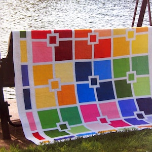 Peut inclure: Une courtepointe colorée avec un motif géométrique de carrés et de rectangles dans des tons de rouge, orange, jaune, vert, bleu et rose. La courtepointe est pliée et drapée sur un banc en bois.