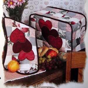 Könnte beinhalten: Ein gestepptes Küchenset in einem rot-weiß-grauen Farbschema. Das Set beinhaltet eine Toasterabdeckung und einen Topflappen, beide mit einem Patchwork-Design mit Herzen. Der Text "Served With Love Kitchen Set" ist oben im Bild gedruckt.