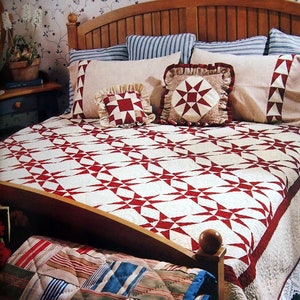 Puede incluir: Una colcha blanca y roja con un patrón de estrellas está sobre una cama con una almohada a rayas blancas, rojas y azules, una almohada blanca con ribetes rojos y azules y una almohada blanca con un patrón de estrellas rojas. La cama tiene una cabecera de madera y una falda de cama blanca.