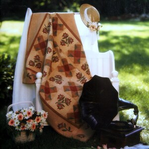 May include: A white wooden bench with a quilt draped over it. The quilt is a patchwork of brown, red, and cream squares with floral designs. A straw hat with a floral embellishment sits on the bench. The bench is in a grassy area with green foliage in the background. The text "Spring Mill Days" is written in brown script on a tan background.