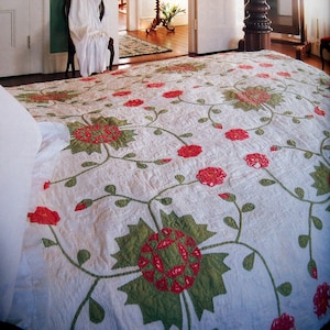 May include: A white quilt with a floral pattern featuring red and green flowers. The quilt is on a bed in a bedroom with white walls and a mirror in the background.