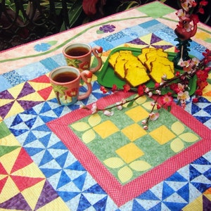 Pode incluir: Uma toalha de mesa de patchwork colorida com um quadrado central verde com formas geométricas amarelas e brancas. Duas chávenas de chá com pires e um prato de bolo de limão estão sobre a mesa. O texto "tea and honey" está escrito em itálico branco sobre um fundo verde.