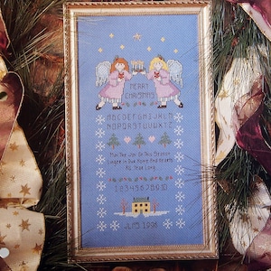 Puede incluir: Patrón de punto de cruz con dos ángeles sosteniendo velas con el texto "Feliz Navidad" y el alfabeto. El patrón también incluye una casa, árboles y el texto "Que la alegría de esta temporada perdure en nuestro hogar y nuestros corazones todo el año".