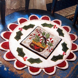 May include: A white felt Christmas-themed rug with a red scalloped border. The center features a square design of a brown pot with green holly and candy canes. The rug is on a blue wooden chair.