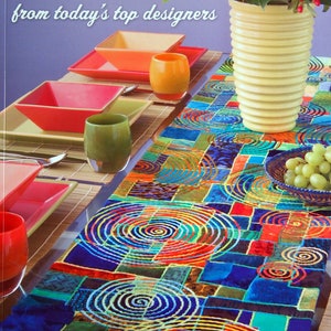 May include: A book cover with the title "Skinny Quilts & Table Runners" and a colourful table runner. The table is set with square plates, glasses, and a vase of flowers. The runner has a geometric design.
