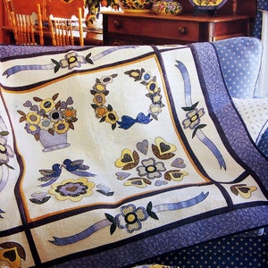May include: A white quilt with a blue and yellow floral design. The quilt is folded and sitting on a blue and white patterned sofa. The quilt features a variety of floral motifs, including wreaths, bouquets, and hearts.
