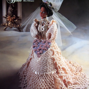 May include: A crocheted wedding dress for a doll, featuring a heart-shaped bouquet and a delicate veil. The dress is a pale pink colour with a lace trim and pearl accents. The doll is wearing a gold necklace and earrings. The image includes the text "The Bridal Belle Collection" and "Miss February".