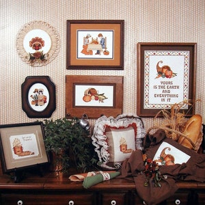 May include: A collection of cross-stitch patterns featuring harvest themes. The patterns include a cornucopia, a basket of pumpkins, a Thanksgiving dinner scene, and a quote about the earth. The patterns are framed and displayed on a brown wooden surface with a checkered tablecloth.