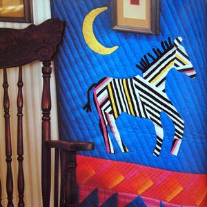 May include: A quilted wall hanging featuring a blue background with a yellow crescent moon and a zebra applique made of black, white, and yellow striped fabric. The bottom section has a red and orange geometric pattern with pink triangular accents.