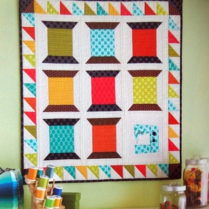 May include: A colorful quilt with a geometric design of spools of thread. The quilt is hanging on a green wall above a white dresser with drawers and shelves. The dresser is decorated with spools of thread, fabric scraps, and other sewing supplies.