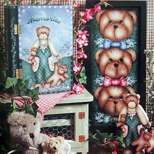 May include: A wooden plaque with a painted image of three teddy bears wearing bows. The plaque is framed in black and has the text "Chezza and Her Bears" and "Teddy Bears Three Plaque" written below the image.