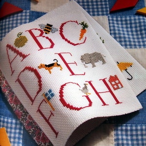 May include: A white fabric alphabet sampler with red embroidered letters A through J. Each letter is decorated with a small embroidered image, such as an apple, bee, carrot, dog, elephant, duck, and house.