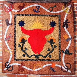 May include: A handmade cowboy quilt with a central design of a red bull skull, surrounded by stars and sunflowers. The quilt features various cowboy-themed appliqués and the text "COWBOY QUILT" on a blue sign.