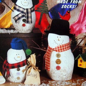 May include: Three snowman door stops made from socks. The snowmen are decorated with plaid scarves, buttons, and twigs for arms. The snowmen are sitting on a wooden surface with a red, blue, and yellow fabric background. The text "A-DOOR-ABLE DOOR STOPS MADE FROM SOCKS!" is printed on the background.