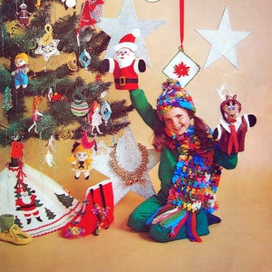 May include: A girl wearing a green sweater and a colorful crocheted scarf holds a reindeer puppet. The girl is sitting in front of a Christmas tree decorated with ornaments, including a Santa Claus puppet, a star, and a snowman. The text "Easy Gifts and Ornaments" is at the top of the image.