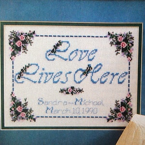May include: Cross-stitch artwork framed in a blue mat and gold frame. The artwork features the words "Love Lives Here" in blue thread, with floral accents. Below the text are the names "Sandra & Michael" and the date "March 10, 1990".