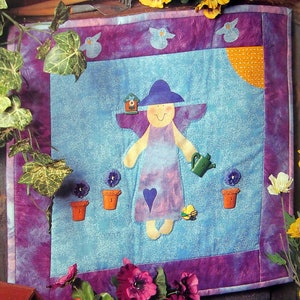 Puede incluir: Una pared acolchada de color morado y azul con un diseño de ángel caprichoso. El ángel lleva un vestido morado y un sombrero azul. El fondo es de color azul claro con nubes blancas. Hay pequeñas macetas y una regadera en la pared acolchada.