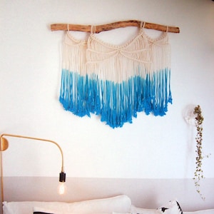 May include: A white macrame wall hanging with blue ombre fringe, hanging from a natural branch, above a bed with striped and patterned pillows.