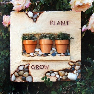 May include: A beige rectangular planter with the words "PLANT" and "GROW" inscribed. The planter has three small terracotta pots with green moss inside, surrounded by small white and brown stones.