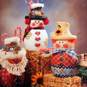 May include: Three handmade Christmas decorations: a snowman with a red and white plaid scarf, a scarecrow with a plaid shirt and a red heart, and a Santa Claus with a white beard and a red and white plaid shirt. All three decorations are sitting on bales of hay.