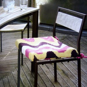 May include: A metal chair with a colorful, patterned cushion. The cushion features a zig-zag design in shades of yellow, purple, pink, and white. The chair is on a wooden deck, with a table and glasses visible in the background.