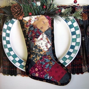 Pode incluir: Uma meia de Natal de patchwork com um padrão floral verde, vermelho e branco. A meia está cheia de um ramo de pinho com bagas vermelhas e uma pinha.
