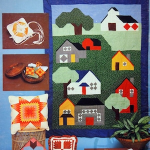 Op de afbeelding: Een kleurrijk quilt met een patchwork-ontwerp met huizen, bomen en een rode schuur. De quilt wordt getoond op een blauwe achtergrond met verschillende gestikte items, waaronder kussens en tafellopers. De tekst "American School of Needlework, excellence in instruction" is zichtbaar in de rechterbenedenhoek.