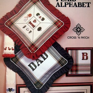 May include: Cross stitch patterns for a plaid alphabet, including the letters B and I, the word "DAD", and the name "MCDANIEL". The patterns are displayed on cushions and framed pieces of fabric.