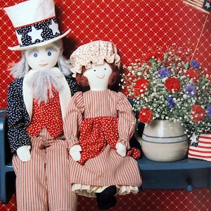 May include: Two fabric dolls sitting on a blue bench. The left doll wears a top hat with white stars on a blue band, a navy blue jacket, and red and white striped pants. The right doll wears a bonnet and a red and white striped dress with a red apron.