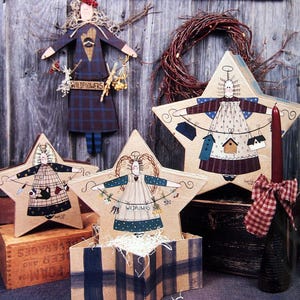 May include: A collection of wooden decor items including a Welcome sign, a hanging figure, and three star-shaped boxes featuring painted figures holding items like birdhouses, watering cans, and wildflowers, with a red candle and plaid ribbon.