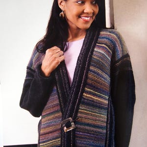 May include: A long-sleeved cardigan featuring a multi-colored horizontal striped pattern in shades of blue, purple, and brown. The garment has solid black sleeves and a black trim along the front opening, secured by a single square-shaped toggle closure.