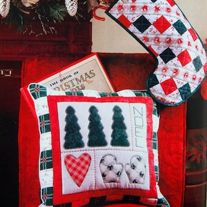 May include: A red and white plaid pillow with a white background and green Christmas trees, a red and white heart, and two white mittens. The pillow is embroidered with the word "Noel". The pillow is sitting on a red chair with a book titled "The Book of Christmas" and a red and green stocking with white trim.