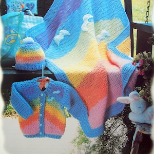 Puede incluir: Una manta de bebé de crochet arcoíris, un jersey de bebé de crochet a juego con botones y un gorro de bebé de crochet. La manta y el jersey tienen un patrón de rayas arcoíris con nubes blancas.