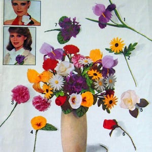 May include: A collection of colorful, handmade paper flowers, including irises, daisies, roses, and carnations, arranged in a beige vase. The flowers are in various colors, such as purple, yellow, red, and white. The image also shows examples of the flowers being worn as corsages.