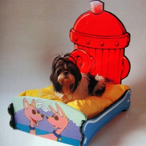 May include: A red fire hydrant shaped dog bed with a yellow cushion. A small black and white dog is lying on the bed. The bed has a blue and purple painted design with two cartoon dogs on the side. The text "SOH NOW" is on the bed.