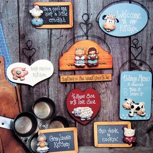 May include: A collection of decorative wooden wall signs featuring painted figures of chickens, cows, sheep, and pigs with text including "I'm a good egg just a little cracked," "Welcome to our Kitchen," "Self service Kitchen," and "Todays Menu."