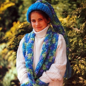 May include: A young person wearing a white sweater, a blue and green crocheted scarf, and a blue crocheted hat with a long tail.