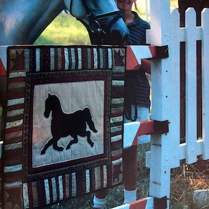 May include: A white horse with a black bridle stands next to a white picket fence. A young person wearing a blue shirt and white shorts is standing next to the horse. A red, white, and blue quilt with a black silhouette of a horse is hanging on the fence.