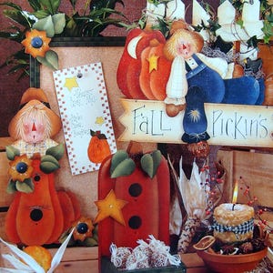 May include: A collection of painted wooden harvest-themed decor featuring scarecrows, pumpkins, and a sign that reads Fall Pickin's. The display includes a vertical sign labeled Harvest Cider Mill, a birdhouse, and a candle in a terracotta bowl.