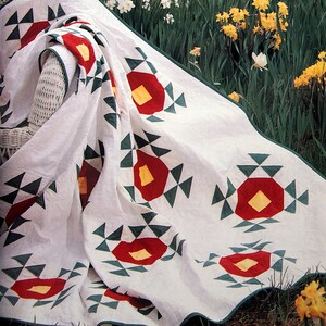 May include: A white quilt with a geometric pattern of red, yellow, and green. The quilt is draped over a white wicker chair in a garden setting with daffodils in bloom.