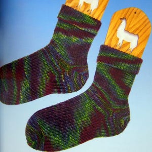 Puede incluir: Un par de calcetines de punto coloridos con un patrón de rayas en tonos verdes, azules y burdeos. Cada calcetín se muestra en una horma de calcetín de madera con un recorte de llama blanco. Fondo azul.