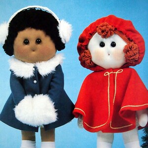 May include: Two handmade dolls wearing winter clothing. The doll on the left has brown skin, black hair, and is wearing a blue coat with white fur trim, white mittens, and black shoes. The doll on the right has white skin, red hair, and is wearing a red hooded cape with gold trim, white socks, and black shoes.