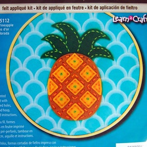 May include: A felt applique kit featuring a golden pineapple with a green leafy top. The kit includes pre-punched felt, thread, a 6-inch wood hoop, a needle, and instructions. Learn a Craft. 72-75112 Golden Pineapple Ananas d'or Piña dorada.