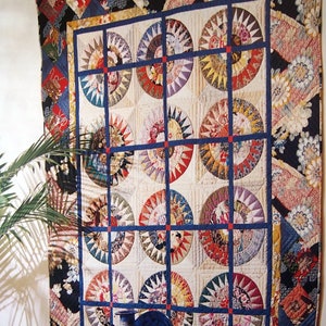 Peut inclure: Une courtepointe colorée avec un motif circulaire, présentant une variété de tissus et de couleurs. La courtepointe est exposée sur un mur, avec une assiette bleue et un bol d'oranges au premier plan.