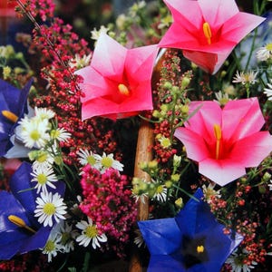 May include: A book cover titled Origami Flowers by Soonboke Smith featuring a vibrant arrangement of folded pink and blue paper flowers interspersed with small white daisies and red heather sprigs, creating a colorful floral display.